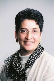 Photo of Pari, a woman with short curly hair, wearing glasses, a white sweater and a dark print scarf. 