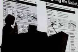 A person is outlined in shadow against a slide showing two ballots
